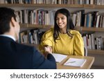 Cheerful Indian student woman talking to male professor in college library, giving handshake to tutor, teacher, thanking for help in study research, sitting at table with bookshelves behind