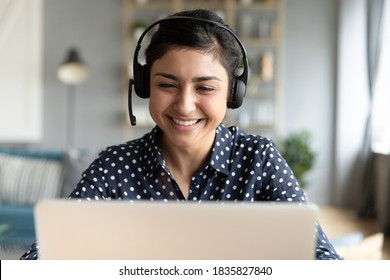 Cheerful Indian woman e tutor sit at desk home office wear headset looks at laptop talk by video call, helpline employee and pleasant chat to client remotely, distance webinar online teaching concept