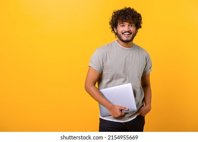 Cheerful Indian Young Man Holding Laptop Standing Isolated On Yellow, Looking At Camera With Happy Smile. Male Student Or Freelancer Carrying Laptop Computer. Copy Space