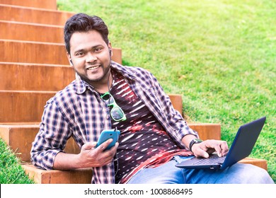 Cheerful indian young man using smart phone and laptop sitting outdoors listening music - Happy asian student holding mobile and pc at park on spring day - Modern concept of multitasking, remote work