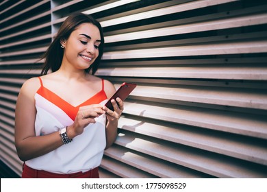 Cheerful influence blogger 20 years old using modern cellular technology for checking web popularity, happy hipster girl with smartphone reading funny publication smiling at urban setting in city