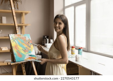 Cheerful inspired young artist woman enjoying creativity  leisure  painting picture in activity studio  drawing at easel and artistic tools  mixing acrylic paints palette  looking at camera