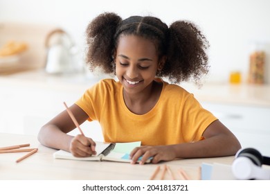 Cheerful inteligent african american teen girl in casual doing homework, sitting at cozy kitchen at home, taking notes or making plan, using pencils and notepad, closeup portrait, copy space