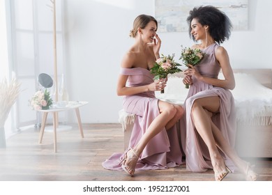 cheerful interracial bridesmaids holding wedding bouquets while talking in bedroom