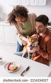 Cheerful Interracial Couple Cuddling Shiba Inu Dog Near Pancakes With Raspberries In Kitchen