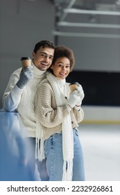 Cheerful interracial couple holding takeaway drink and looking at camera on ice rink  - Shutterstock ID 2232926861