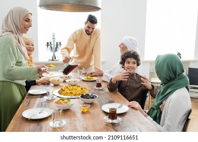 Cheerful interracial family serving pilaf and pouring tea near dinner at home