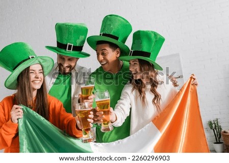 cheerful and interracial friends in green hats holding glasses of beer and Irish flag while celebrating Saint Patrick Day 