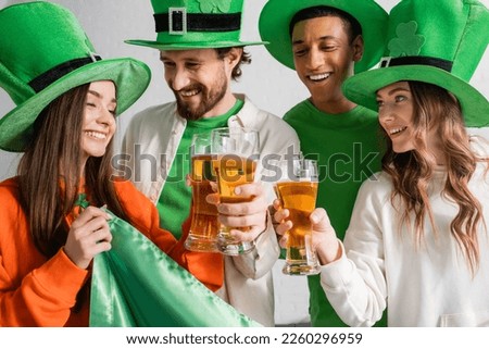 cheerful and interracial friends in green hats clinking glasses of beer near Irish flag while celebrating Saint Patrick Day 