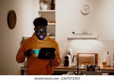 A cheerful interracial guy is standing at his cozy loft and having a video call on a tablet at night while drinking and enjoying tea. Online date concept. Happy man flirting while having a video call - Powered by Shutterstock