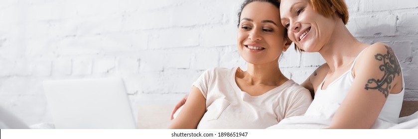 Cheerful Interracial Lesbian Couple Watching Movie On Computer In Bed, Banner