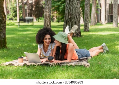 Cheerful Interracial Same Sex Couple With Coffee To Go Using Devices On Blanket In Park