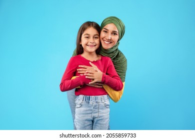 Cheerful Islamic Mother Wearing Hijab Hugging Her Little Daughter Smiling To Camera Posing On Blue Background. Arabic Mom And Her Kid Girl Standing And Embracing Together In Studio