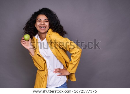 Cheerful joyful funny dark-skinned young lady, with curly hair, in a white T-shirt and a coat, holds a green apple in his hand and laughs, looks straight, stands on a gray background. Copy space.