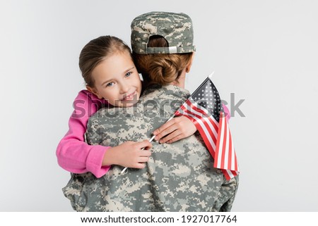 Cheerful kid with american flag embracing mother in military uniform isolated on grey 