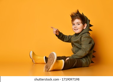 Cheerful kid boy in dark green hoodie with dinosaurs spikes at his back and cowl and rubber boots sits on the floor pointing finger at upper corner at free copy space