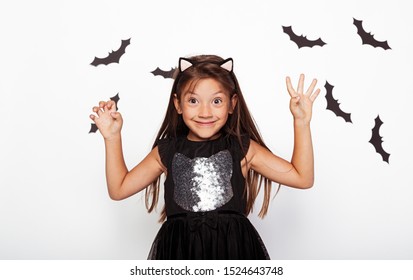 Cheerful Kid In Cat Costume Standing With Raised Hands On White Background With Flying Black Bats And Looking At Camera