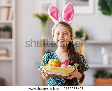 Cheerful kid  girl in casual clothes and bunny ears on head smiling and looking at camera while standing in a cozy living room during preparation for the Easter holiday  with basket of colorful eggs