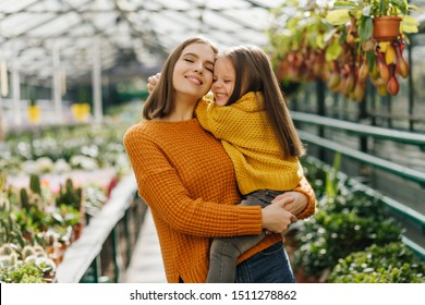 Cheerful kid in yellow sweater sitting on mother's hands. Photo of blissful family posing in greenhouse. - Powered by Shutterstock