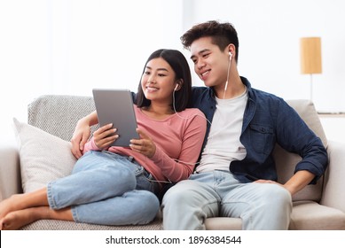Cheerful Korean Couple Using Digital Tablet Computer Watching Movie Relaxing On Day-Off Sitting On Sofa At Home. Asian Family Browsing Internet Together In Living Room On Weekend