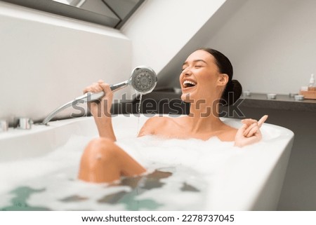 Cheerful Lady Having Fun Taking Bath And Singing Holding Shower Head Like Microphone Sitting In Water With Foam Bubbles In Modern Bathroom At Home. Beauty And Relaxation Leisure