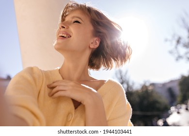 Cheerful lady making selfie at sunny day. Pretty young girl with short dark hair, yellow clothes and nude makeup, smiling and narrowing eyes while waving head