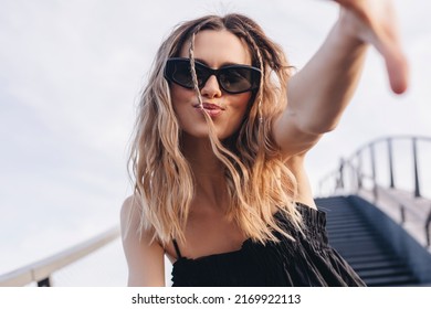 Cheerful lady making selfie at sunny day. Pretty young girl with curly blonde hair and two thin braids towards to camera, smiling and wear black glasses and dress, send blow kiss under sky and stairs. - Powered by Shutterstock