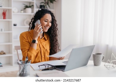 Cheerful Lady Talking On Cellphone Holding Paper Sitting At Desk And Using Laptop Computer Working Online In Modern Office. Entrepreneurship And Business Career, Distance Freelance Job Concept