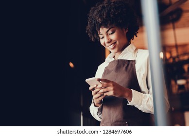Cheerful lady waitress having break from work. She holding mobile phone in hands and sending message to her friends