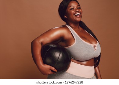 Cheerful large african woman with a medicine ball on brown background. Plus size female in sports clothing looking at camera and laughing.