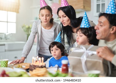 Cheerful Latin American Family Wearing Birthday Caps, Blowing Candles On A Cake While Celebrating Birthday Together At Home