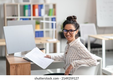 Cheerful latin female general manager checking reports in modern office, sitting at workplace with computer and smiling at camera, copy space