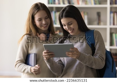 Cheerful Latin student girl showing learning online presentation on tablet to classmate, holding digital gadget, looking at screen, smiling, using device in college, school library