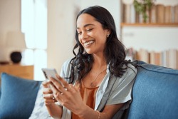 Cheerful Latin Woman Using Smartphone During Video Call While Sitting On Sofa At Home. Smiling Hispanic Girl Chatting On Social Network On Smart Phone. Mixed Race Girl Laughing While Watching Video.