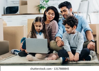 Cheerful Latino Family Looking At Laptop While Sitting On Carpet In New Home