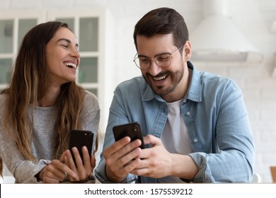 Cheerful Laughing Millennial Couple Using Smart Phones Play Mobile Games Together, Happy Girl And Guy Friends Or Family Customers Having Fun With Cellphones Devices Enjoy Buying Online Sit At Table