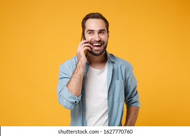 Cheerful Laughing Young Man In Casual Blue Shirt Posing Isolated On Yellow Orange Background, Studio Portrait. People Sincere Emotions Lifestyle Concept. Mock Up Copy Space. Talking On Mobile Phone