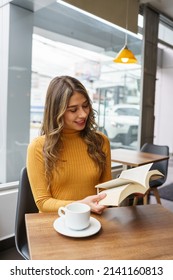 cheerful lifestyle of attractive young latin woman with long blonde hair, enjoying reading a book, at the table a cup of coffee, intellectual pastime, knowledge and learning