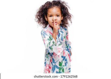 Cheerful little african girl wearing white floral dress standing isolated over white background, showng silence gesture
