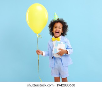 Cheerful little black boy with curly hair in stylish suit and party hat smiling while standing against blue background  with yellow balloon and present in hands