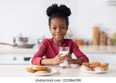 Cheerful Little Black Girl Sitting At Table, Enjoying Milk And Cookies, Cute African American Child Having Snacks In Kitchen, Having A Bite With Healthy Calcium Drink And Homemade Pastry