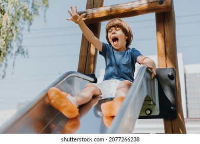 cheerful little boy in a blue T-shirt and white shorts rolls down a metal slide in city park. child walks outdoors. healthy active recreation. lifestyle. space for text. High quality photo - Powered by Shutterstock