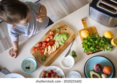 Cheerful little boy cooking fresh healthy breakfast at kitchen with avocado, cottage cheese, fruit and berries. Funny male kid trying eating blueberry ingredient at cuisine preparing sandwich
