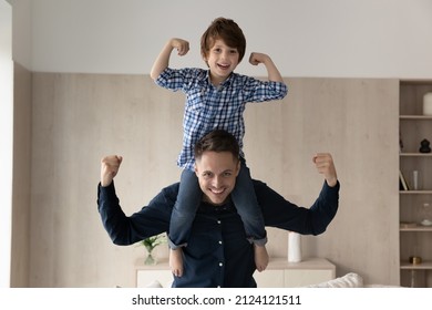 Cheerful little boy riding on happy dads neck and shoulders, making powerful hands. Strong daddy keeping balance with kid on top, flexing arm muscles, smiling at camera. Father and son portrait
