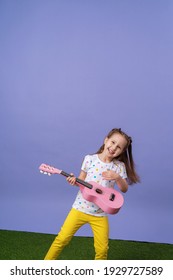 cheerful little Caucasian girl of 4 years old in fashionable bright clothes plays a pink ukulele guitar. The child plays a musical instrument and sings on a lilac background and green grass.