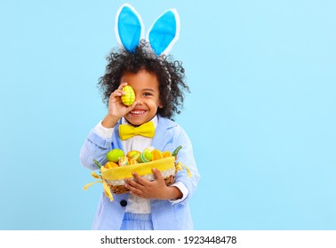 Cheerful little ethnic boy with Afro hair in Easter costume and bunny ears smiling while covering eye with colorful egg against blue background