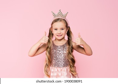 cheerful little girl in dress and crown showing thumbs up isolated on pink