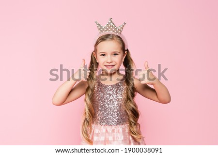 cheerful little girl in dress and crown showing thumbs up isolated on pink