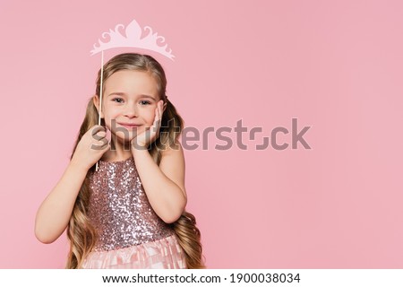 cheerful little girl in dress holding carton crown on stick above head isolated on pink