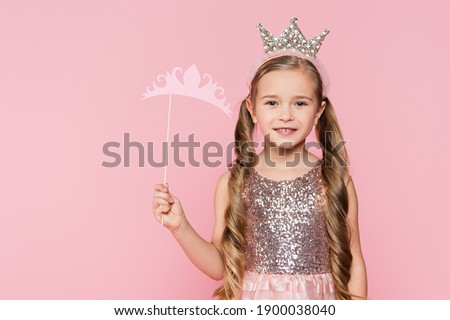 cheerful little girl in dress holding carton crown on stick isolated on pink